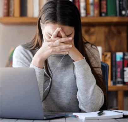A woman sitting at a library table in front of a laptop, holding her head in her hands