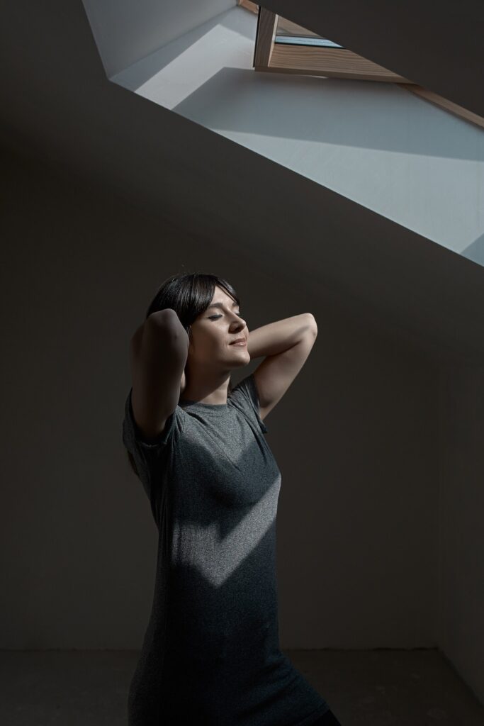 Woman alone in a room with her arms up and eyes closed, standing in slight through a window